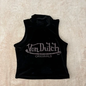 Svart ärmlös polotopp från Von Dutch - Snygg svart ärmlös croppad topp med polokrage från märket Von Dutch i mjukt sammetstyg. Toppen har ett glittrigt rosa Von Dutch-logotyp framtill. 