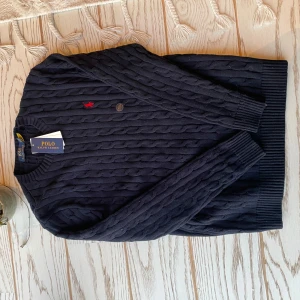 Svart stickad tröja från Ralph Lauren - Säljer en stilren svart stickad tröja från Ralph Lauren med kabelstickat mönster. Tröjan har en klassisk rund hals och långa ärmar. Den röda logotypen på bröstet ger en snygg kontrast. Perfekt för kyligare dagar!