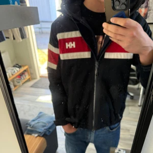 Svart vindjacka från Helly Hansen - Snygg svart vindjacka från Helly Hansen med röda och vita detaljer. Jackan har en dragkedja framtill och långa ärmar. Perfekt för blåsiga dagar.