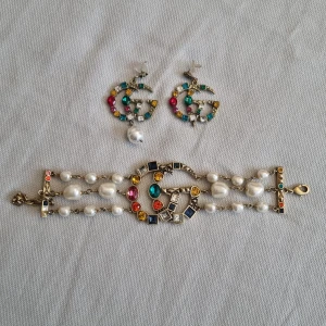 Färgglatt armband och örhängen med pärlor - "Gucci" set med armband och örhängen dekorerade med färgglada stenar och pärlor. Armbandet har en mix av blå, röda och gula stenar samt vita pärlor. Örhängena matchar med liknande stenar och en hängande pärla. Aldrig använt. 