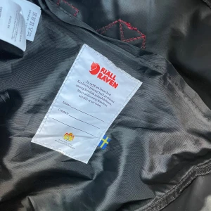 Svart ryggsäck från Fjällräven - Säljer en klassisk svart Fjällräven Kånken ryggsäck med vinröda axelremmar och handtag. Ryggsäcken har en framficka med dragkedja och det ikoniska Fjällräven-märket på framsidan. Perfekt för vardagsbruk och utflykter. Något dammig på bild men i övrigt nyskick.