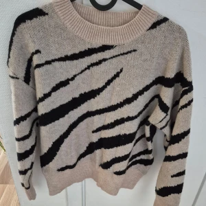 Beige och svart stickad tröja - Säljer en mysig stickad tröja i beige med svarta zebramönster. Tröjan har långa ärmar och en rund halsringning. Perfekt för kyliga dagar!