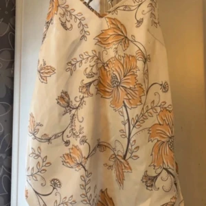 Blommig topp  - Säljer en snygg topp  med blommigt mönster i beige och orange. Toppen  har en unik ringdetalj i ryggen och en v-ringad hals. Perfekt för sommarens varma dagar!