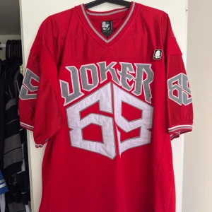 Joker brand jersey  - Säljer en cool röd jersey  med stort 'Joker 69' tryck i grått och vitt. Tröjan har en v-ringad hals och korta ärmar med randiga detaljer. Perfekt för dig som vill sticka ut!