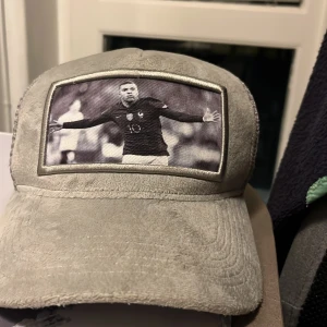 Grå keps från Evo Sportswear med Mbappé  - Snygg grå keps från Evo Sportswear med ett svartvitt foto på framsidan. Kepsen har en klassisk design med böjd skärm och justerbar passform. Perfekt för sportiga tillfällen eller som en stilren accessoar.