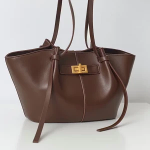 Brun axelväska med gulddetalj - Snygg brun axelväska i skinn med elegant gulddetalj på framsidan. Väskan har en stilren design med långa axelremmar och en praktisk stängning. Perfekt för att bära dina dagliga nödvändigheter med stil.