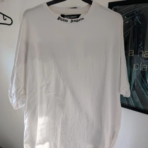 Vit t-shirt från Palm Angels - Säljer en stilren vit t-shirt från Palm Angels med deras ikoniska logga tryckt i svart och rosa på baksidan. T-shirten har en normal passform och är kortärmad, perfekt för en avslappnad look.