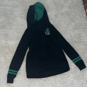 Svart hoodie med gröna detaljer - Säljer en svart Harry Potter hoodie med gröna ränder på ärmarna och en grön insida av huvan. Den har en dragkedja framtill och ett emblem på bröstet. Perfekt för en avslappnad stil. Ny pris: 700