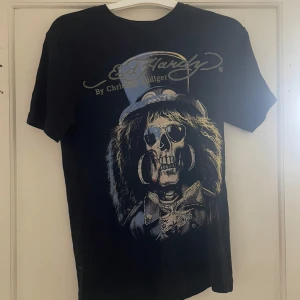 Svart t-shirt från Ed Hardy - Säljer en svart t-shirt från Ed Hardy med ett coolt tryck av en dödskalle med hatt och solglasögon. T-shirten har korta ärmar och är perfekt för dig som gillar en edgy stil. Passar bra till jeans eller shorts för en avslappnad look.