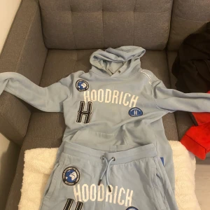 Ljusblå dress från Hoodrich - Säljer en ljusblå hoodie från Hoodrich med stora tryck och logotyper i vitt och svart. Hoodien har en klassisk design med känguruficka och justerbar huva. Perfekt för en avslappnad stil.