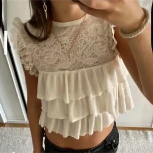 Beige topp med spetsdetaljer från Zara - Säljer en söt beige topp från Zara med spetsdetaljer och volanger. Toppen har ett vackert blommönster på framsidan och är ärmlös med spetskantade axlar. Perfekt för en romantisk look.