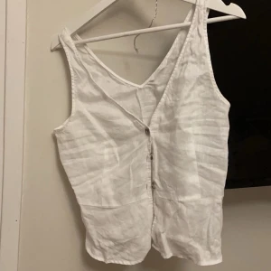 Vit ärmlös linnetopp - Säljer en stilren vit linnetopp med knappar framtill. Toppen är ärmlös och har en v-ringad design, perfekt för varma dagar. står ej strl, är från hm, passar mig bra som är en S 