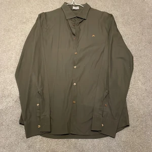 Olivgrön skjorta från J.Lindeberg - Snygg olivgrön skjorta från J.Lindeberg med klassisk krage och knappar framtill. Skjortan har en diskret broderad logga på bröstet och är tillverkad i ett mjukt material. Perfekt för en stilren look.