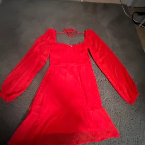 Röd klänning med puffärmar - Snygg röd klänning med puffiga långärmar och ett diskret prickigt mönster. Klänningen har en knytning vid halsen och en volangdetalj vid fållen. Perfekt för en färgstark look!