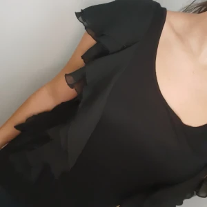 Topp med volangdetaljer - Snygg svart kortärmad tröja med volangdetaljer längst längden. Perfekt för en stilren look. Tröjan har en rund halsringning och är tillverkad i ett lätt material som ger en fin passform. Storlek M från H&M.