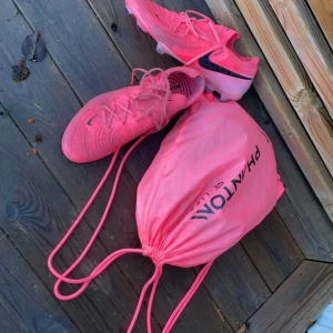 Nike phantom GX || Elite FG Mad Brilliance - Snygga rosa fotbollsskor från Nike med svart logga och snörning. Skorna har en modern design och kommer med en matchande rosa påse. Perfekta för fotbollsplanen! Använda 3-4 ggr, 2800kr nypris
