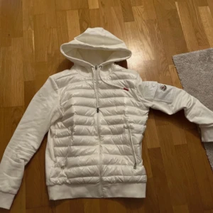 Vit pufferjacka med huva - Snygg vit pufferjacka med huva och dragkedja framtill. Jackan har långa ärmar och en quiltad design. Perfekt för kyligare dagar.