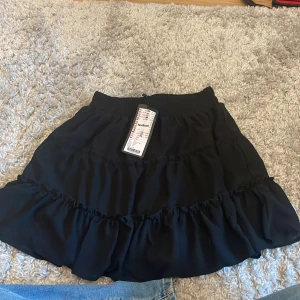 Svart kjol från New Yorker - Snygg svart kjol från New Yorker med volangdetaljer. Perfekt för att ge en trendig touch till din outfit. Kjolen har en elastisk midja för extra komfort. Aldrig använt men e kanske ett år gammal 