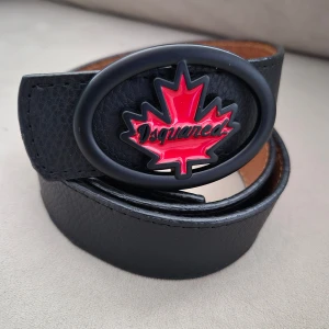 Svart bälte med röd lönnlövsdetalj - Snyggt svart bälte från Dsquared med en cool oval spänne prydd med en röd lönnlövsdetalj. Perfekt för att ge din outfit en unik touch. Tillverkat i skinn för en hållbar och stilren look.