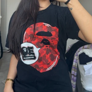 Äkta Bape T-shirt med röd kamouflagemönster - Snygg svart t-shirt med ett stort rött kamouflagemönster på framsidan. T-shirten har korta ärmar och en cool design som sticker ut. Perfekt för en avslappnad stil. Äkta bape