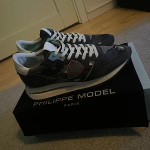 sneakers från Philippe Model - Snygga sneakers från Philippe Model i en cool grå och svart camouflage-design. Skorna har vita snören och en vit sula som ger en fräsch look. Perfekta för dig som vill ha en stilren men ändå unik stil.