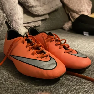 Orange fotbollsskor från Nike - Säljer ett par orangea fotbollsskor från Nike med svarta och silverfärgade detaljer. Skorna har snörning och är designade för att ge bra grepp på planen. Perfekta för fotbollsträning och matcher.