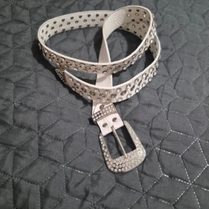 Vitt bälte med strass - Snyggt vitt bälte dekorerat med silverfärgade nitar och strassdetaljer. Bältet har en glittrig spänne som ger en extra touch av stil. Perfekt för att lyfta vilken outfit som helst. Använd en gång längd 110 bred 3cm.