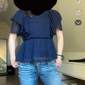 Mörkblå blus med volangdetaljer - Säljer en söt mörkblå blus med volangdetaljer och prickigt mönster. Blusen har korta ärmar och en luftig passform, perfekt för en stilren look. Passar bra till jeans eller kjol.