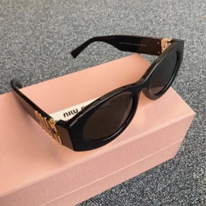 Miu Miu solglasögon  - Snygga svarta solglasögon från Miu Miu med rundade linser och guldiga detaljer på skalmarna. Perfekta för en stilren look. Kommer med ett rosa fodral och matchande låda.