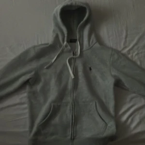 Grå hoodie från Polo Ralph Lauren - Säljer en stilren grå hoodie från Polo Ralph Lauren med dragkedja och klassisk logga på bröstet. Perfekt för en avslappnad look. Den har en justerbar huva med snören och praktiska fickor framtill.