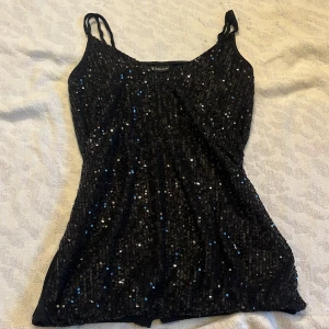 svart glitter linne - Säljer ett svart linne från Bodyflirt med glittrande paljetter på framsidan. Linnet har smala axelband och en enkel design på baksidan. Perfekt för en kväll ute eller när du vill glittra lite extra. Har ett litet märke på baksidan längst ner men inget man märker av. storlek 32/34