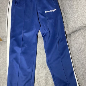 Blå träningsbyxor från Palm Angels - Blå Palm Angels byxor i storlek L .Byxorna har en elastisk midja och vita ränder längs sidan. Om du har några frågor?? Tveka inte på att höra av dig!