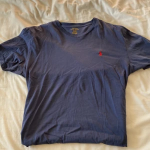 Blå t-shirt från Polo Ralph Lauren - Säljer en klassisk blå t-shirt från Polo Ralph Lauren med röd broderad logga på bröstet. T-shirten är kortärmad och tillverkad i mjuk bomull. Perfekt för en avslappnad stil.