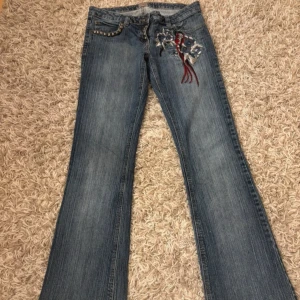 Blå jeans med broderade detaljer - Snygga blå jeans med unika broderade blommor och fåglar i rött och vitt. Byxorna har en låg midja och en bootcut-stil. Midjebredden: 40/41*2 cm Lårbredden: 25 cm Bredden längst ner: 23 cm Innerbenslängden: 82 cm 💖💖💖