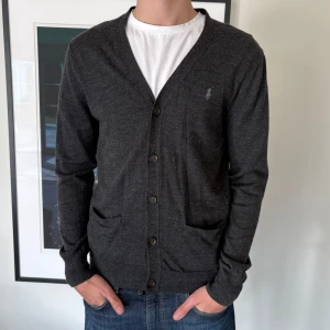 100 % Merinoull cardigan från Ralph Lauren - Tja. En stilren mörkgrå cardigan från Ralph Lauren i merinoull. Koftan har en slim fit passform med knappar framtill och två fickor. Perfekt att ha till sommaren🌟priset går o diskutera, vid frågor hojta! Den är i helt nytt skick!