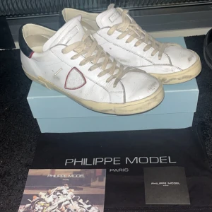 Vita sneakers från Philippe Model - Säljer ett par riktigt snygga  vita sneakers från Philippe Model med en röd detalj på sidan och svart häl. Skorna har snörning och en rund tå. Perfekta för en stilren look nu till våren och sommaren. Skorna är i strl 45 och är sparsamt använda och är i bra skick (slitningarna och smutsen tillhör skon de är inte slitna), köpta på Philippe Models hemsida (kvitto finns) Ny pris 4599 mitt pris 1250, kan gå ner i pris vid en schysst och snabb affär!!