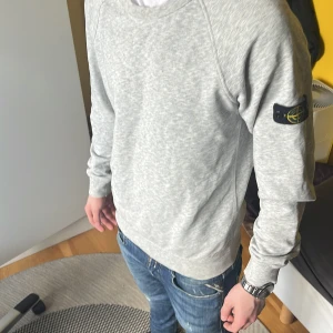 Grå sweatshirt från Stone Island - Säljer en stilren grå sweatshirt från Stone Island. Tröjan har långa ärmar och en rund halsringning. Den är tillverkad i ett mjukt material och har en ikonisk logga på ärmen. Perfekt för en avslappnad stil.