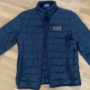 Svart quiltad jacka från Emporio Armani - Snygg svart quiltad jacka från Emporio Armani EA7. Jackan har en dragkedja framtill och är perfekt för kyligare dagar. Den har en stilren design med märkets logga på bröstet.