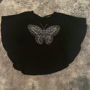 Svart topp med fjärilsdetalj - Snygg svart topp med en glittrig fjäril på framsidan. Toppen har en lös passform och korta ärmar, väldigt mjukt material. Den har ett fint knyte på ryggen. Den kommer från Lindex och är i storleken 146-152. 