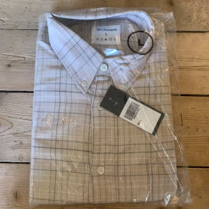 Beige rutig skjorta - Säljer en beige rutig skjorta i 100% bomull. Skjortan har en klassisk krage och knappar framtill. Perfekt för en stilren look. Aldrig använd.