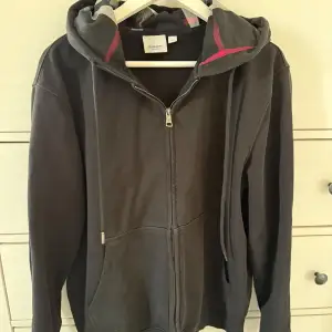 Säljer en stilren svart hoodie från Burberry med dragkedja och huva. Den har en klassisk design med snörning i huvan och en diskret logga. Perfekt för en avslappnad look.