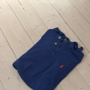 Blå pikétröja från Ralph lauren  - Säljer en klassisk blå pikétröja från Ralph Lauren med en liten röd logga på bröstet. Tröjan har korta ärmar och knappar vid kragen. Perfekt för en stilren och avslappnad look.