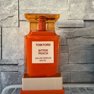 Tom Ford Bitter Peach Eau de Parfum - Säljer en exklusiv Tom Ford Bitter Peach Eau de Parfum i en elegant orange flaska på 100 ml. Flaskan har en fyrkantig form med en stilren etikett och ett matchande lock. Perfekt för den som älskar fruktiga och lyxiga dofter.