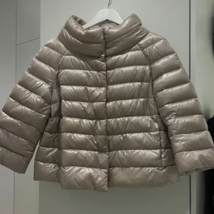 Herno Sofia stl 3 - Sand/beige dunjacka från Herno med en glansig finish. Jackan har en hög krage och trekvartsärmar,  quiltad och fylld med ultralätt gåsdun. Stl 3 motsvarar M/L