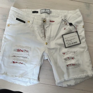 Vita shorts från Philipp Plein - Snygga vita shorts från Philipp Plein med fransiga detaljer pärlor i rött och silver. Shortsen har en dragkedja och knappar i guld och är helt nya! 