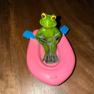 Grön groda i rosa båt - Charmig grön groda som sitter i en rosa båt med blå åror. Perfekt som dekoration eller leksak för barn. Den är i plast så skulle nog tro att den inte går sönder så lätt! köpt second hand❤️