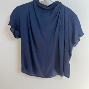 Blå blus från Zara - Säljer en elegant blå blus från Zara med korta ärmar och en draperad hals. Perfekt för en stilren look. Blusen har en knappdetalj i nacken för enkel påklädning.