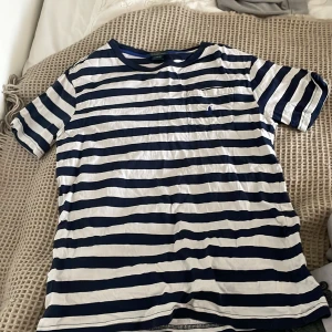 Randig t-shirt från Polo Ralph Lauren - Säljer en snygg randig t-shirt från Polo Ralph Lauren i blått och vitt. T-shirten har en bröstficka med en broderad logotyp. Perfekt för en avslappnad stil!