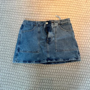 Blå jeanskjol med fickor - Snygg blå jeanskjol med två stora framfickor och knappstängning. Perfekt för en avslappnad stil. Passar bra till en enkel t-shirt eller blus.