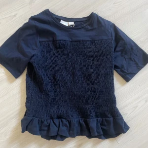 Smockad topp från Gina Tricot - Säljer en mörkblå topp från Gina Tricot med korta ärmar och volangdetalj vid nederkanten. Toppen har en smockad textur på framsidan som ger en fin effekt. Perfekt för en stilren look!